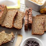 Sliced loaf of grain-free banana bread with chocolate chips decorating the top. A slice is off to the left side, spread with seed butter.