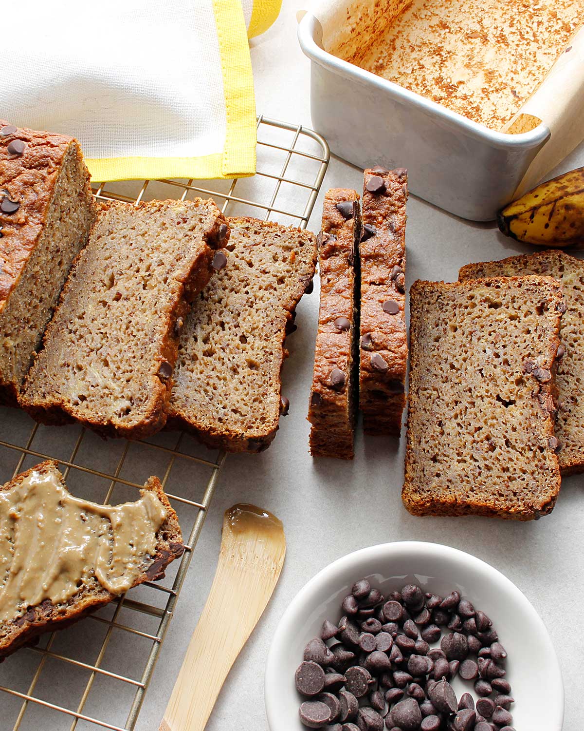 Sliced loaf of grain-free banana bread with chocolate chips decorating the top. A slice is off to the left side, spread with seed butter.