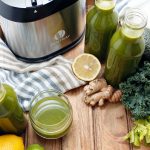 Green juice in glass and bottles with juicer in background.